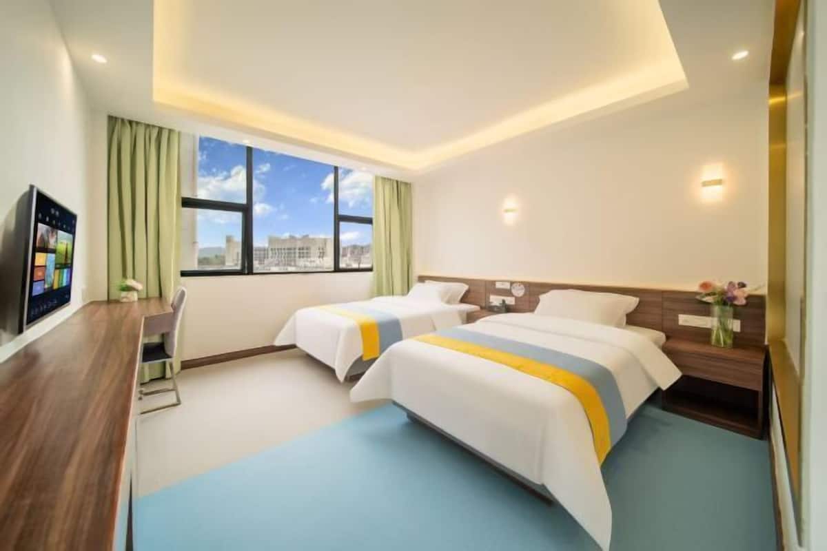 A hotel room with two beds, a desk, a TV, a window with a city view, and a nightstand with a vase of flowers.
