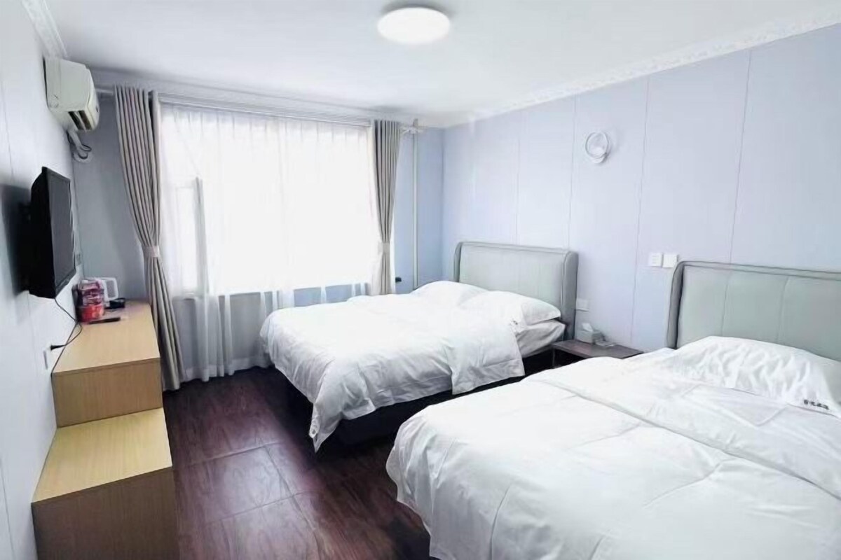 A hotel room with two beds, a flat-screen TV, a small table, and a window with curtains.