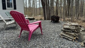 A red Adirondack chair in front of a white house with black shutters, surrounded by a gravel area and a pile of logs. - Home Away from Home- Discover Comfort and Convenience in this 4 Bedroom Home  (Holden)