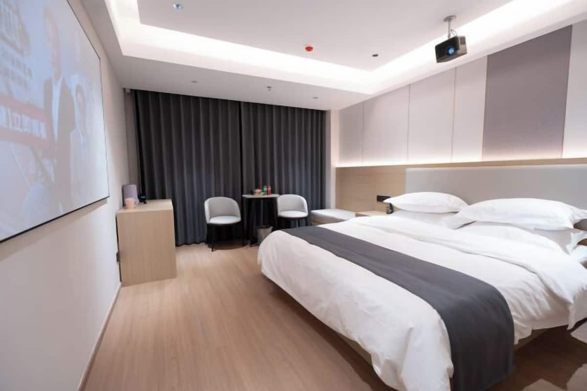 A hotel room with a large bed, a desk, two chairs, a projector screen, and a ceiling-mounted projector.