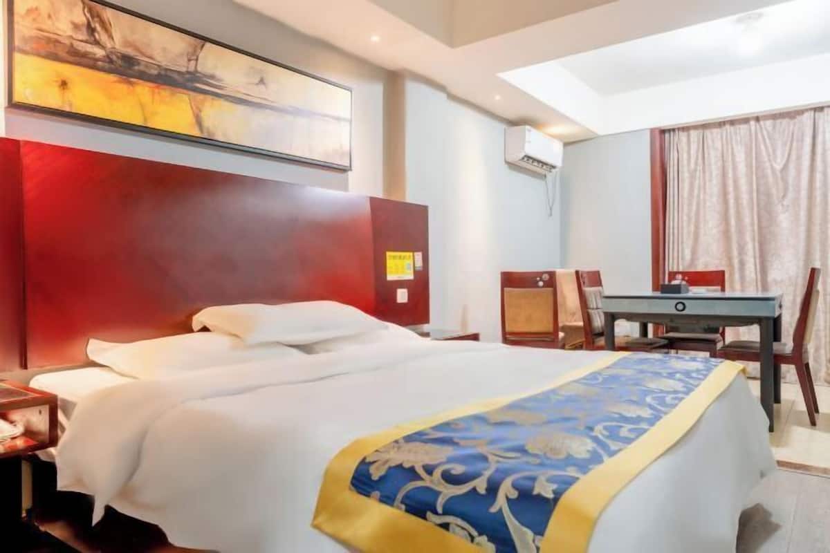 A hotel room with a large bed, a desk, and a chair. There is a painting on the wall and an air conditioning unit above the desk.