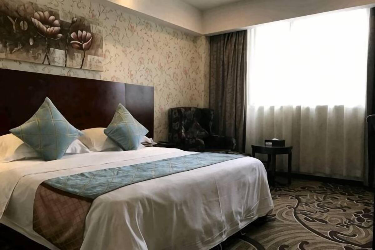 A hotel room with a large bed, a headboard, a chair, a small table, and a window with curtains.