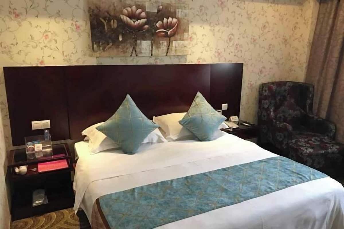 A hotel room with a large bed, two pillows, a nightstand with a phone and a lamp, a chair, and a floral wallpaper.