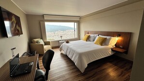 A hotel room with a bed, a desk with a laptop, a chair, a nightstand with a lamp, and a window with a city view. - Di Carbone Hotel Gastrobar (Facatativá)