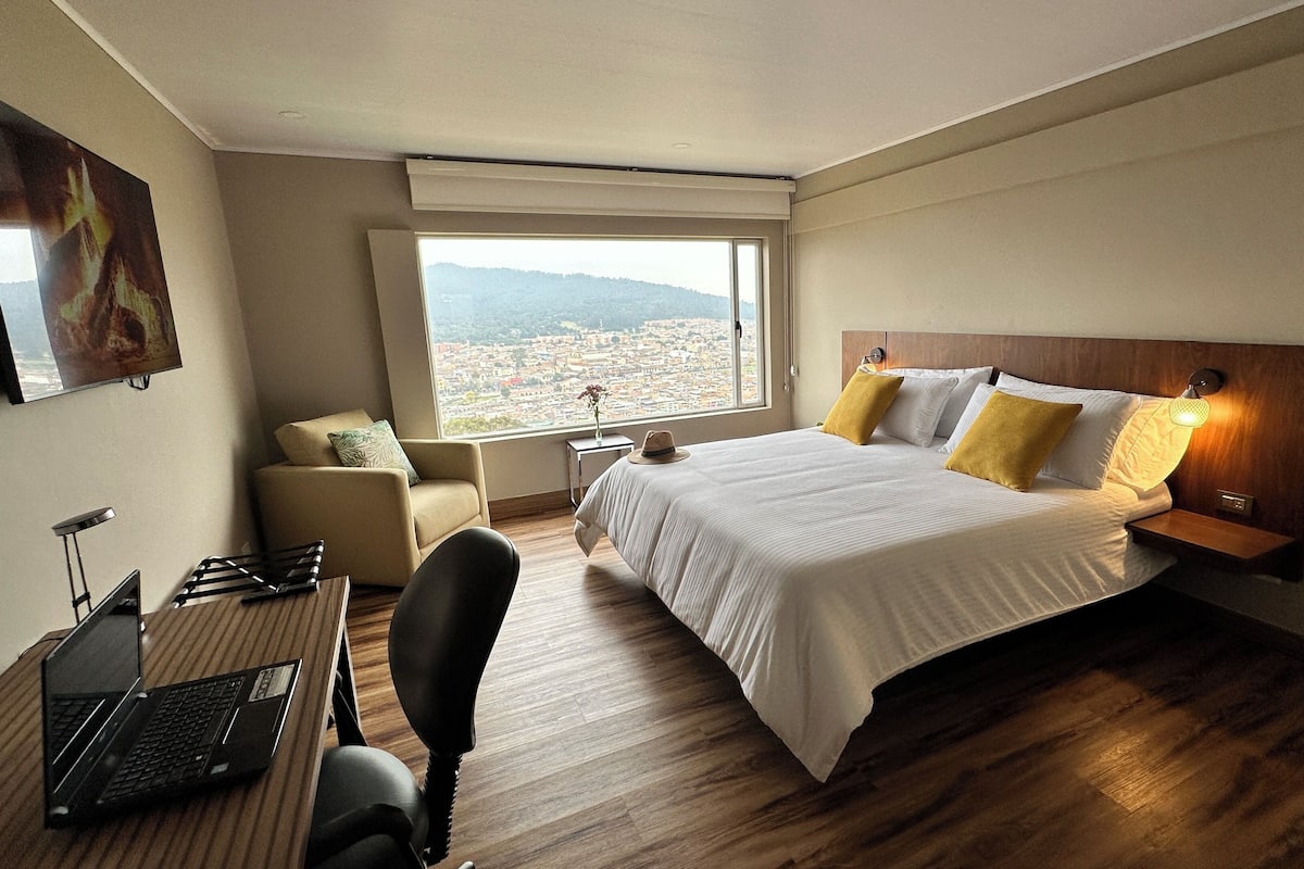 Une chambre d’hôtel comprenant un lit, un bureau avec un ordinateur portable, une chaise, une table de chevet avec une lampe et une fenêtre donnant sur la ville.