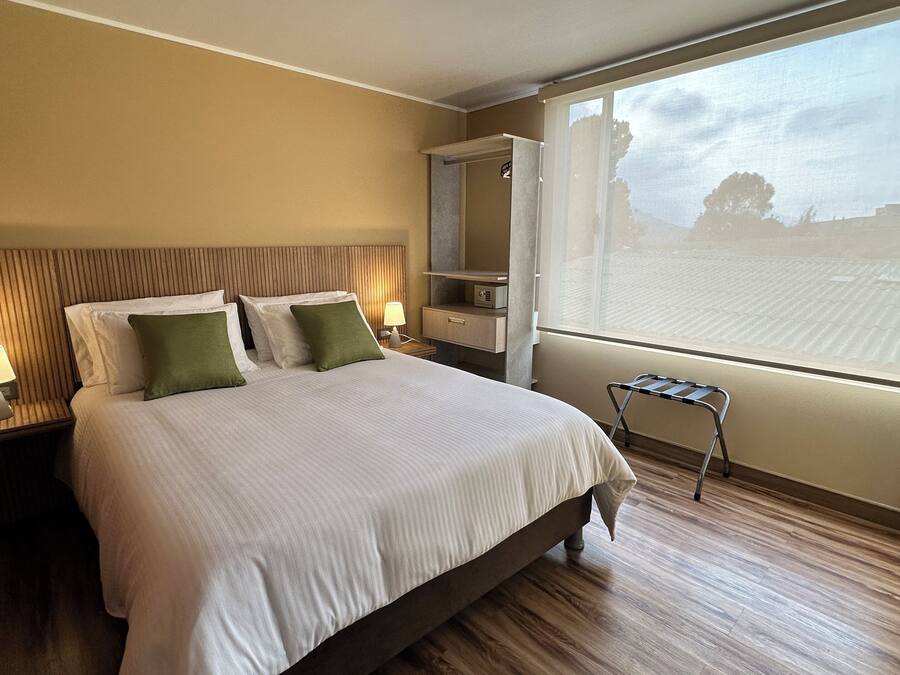Habitació d'hotel amb un llit gran, dues taules de nit amb làmpades, un petit tamboret, un armari i una finestra gran amb vista al cel i als arbres.
