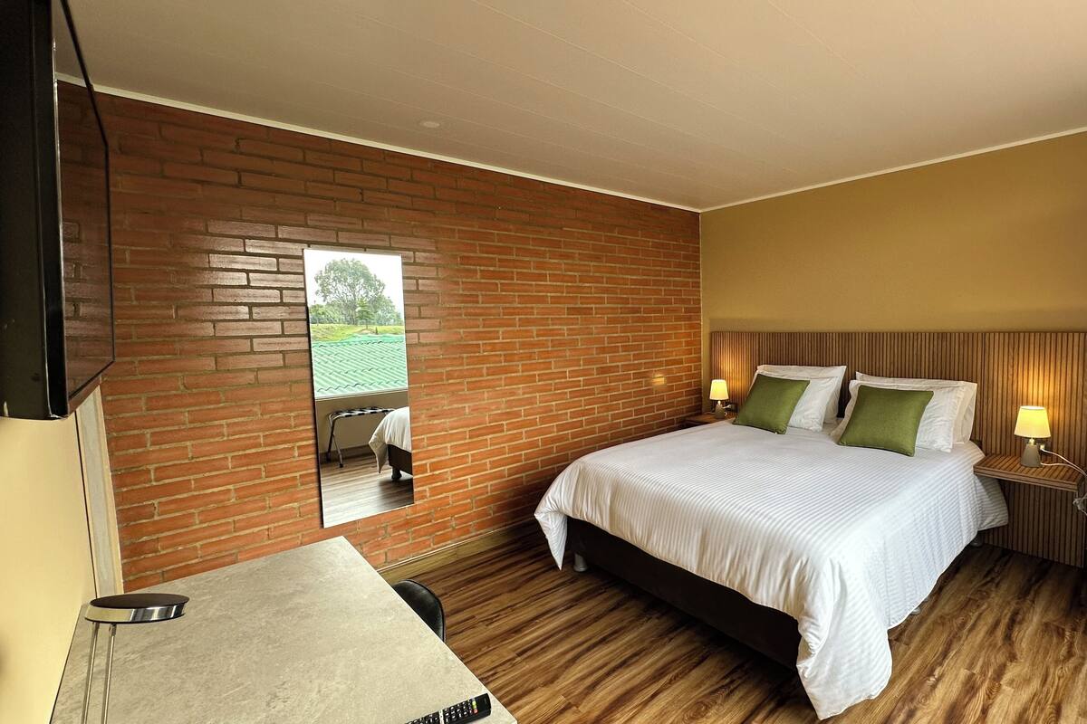 Une chambre d’hôtel avec un mur en briques, un lit avec une literie blanche et des oreillers verts, une table de chevet avec une lampe, et une petite table avec une télécommande.