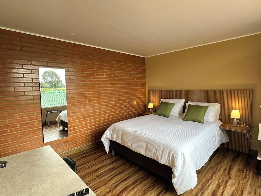 A hotel room with a brick wall, a bed with white bedding and green pillows, a bedside table with a lamp, and a small table with a remote control.