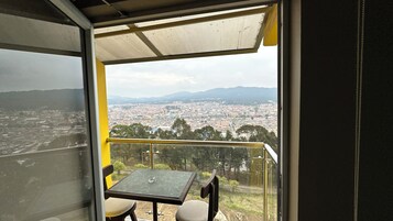 A balcony with a table and chairs, overlooking a cityscape with buildings and mountains in the distance.