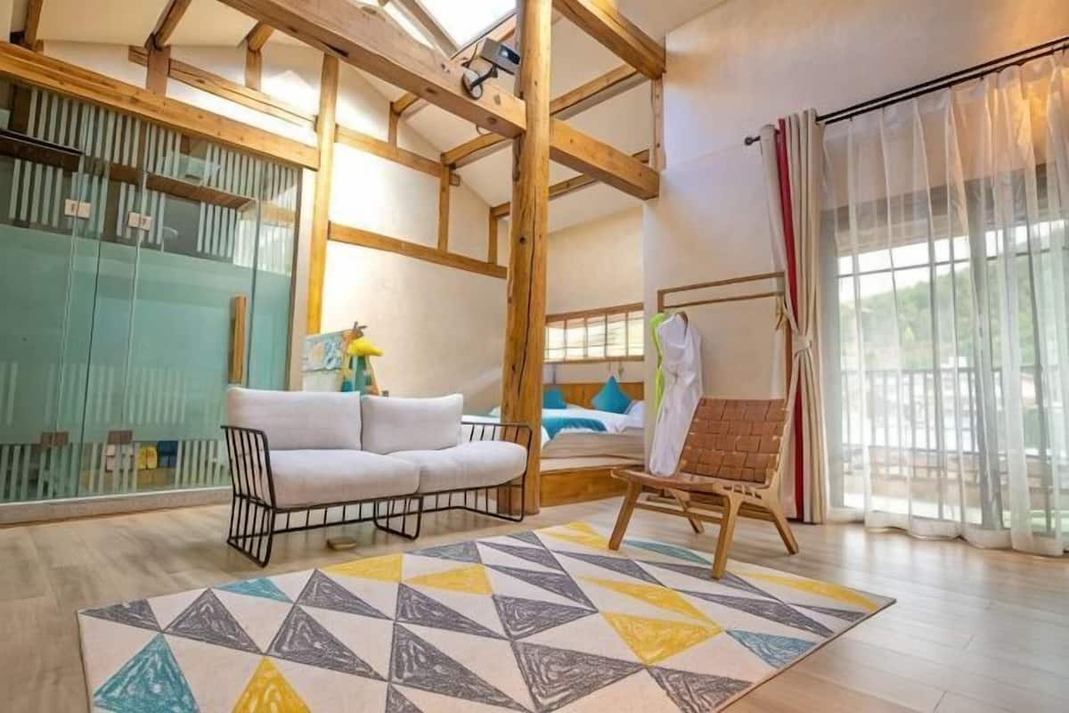 Una habitación con techo de madera, un sofá, una silla, un tapete estampado y un ventanal con cortinas.