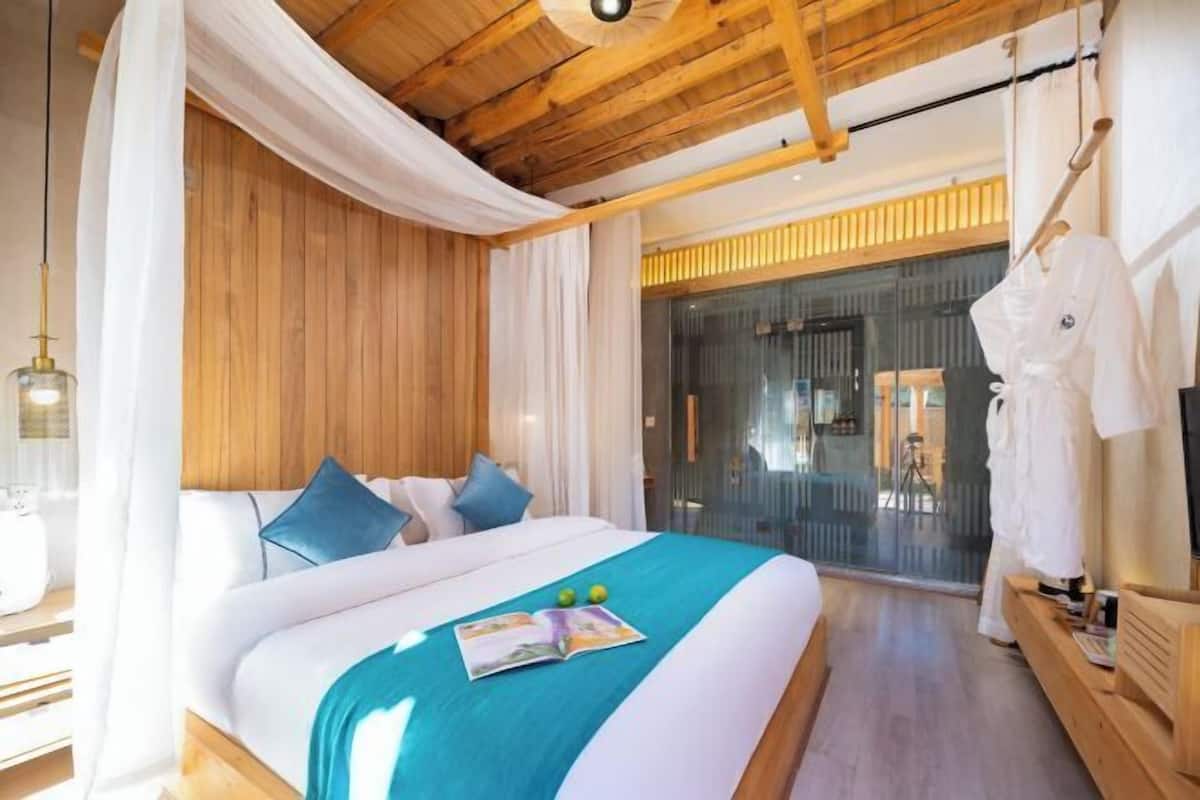 Una habitación de hotel moderna con techo de madera, una cama grande con cubrecama color turquesa, cortinas blancas y una puerta de vidrio que da a un balcón.