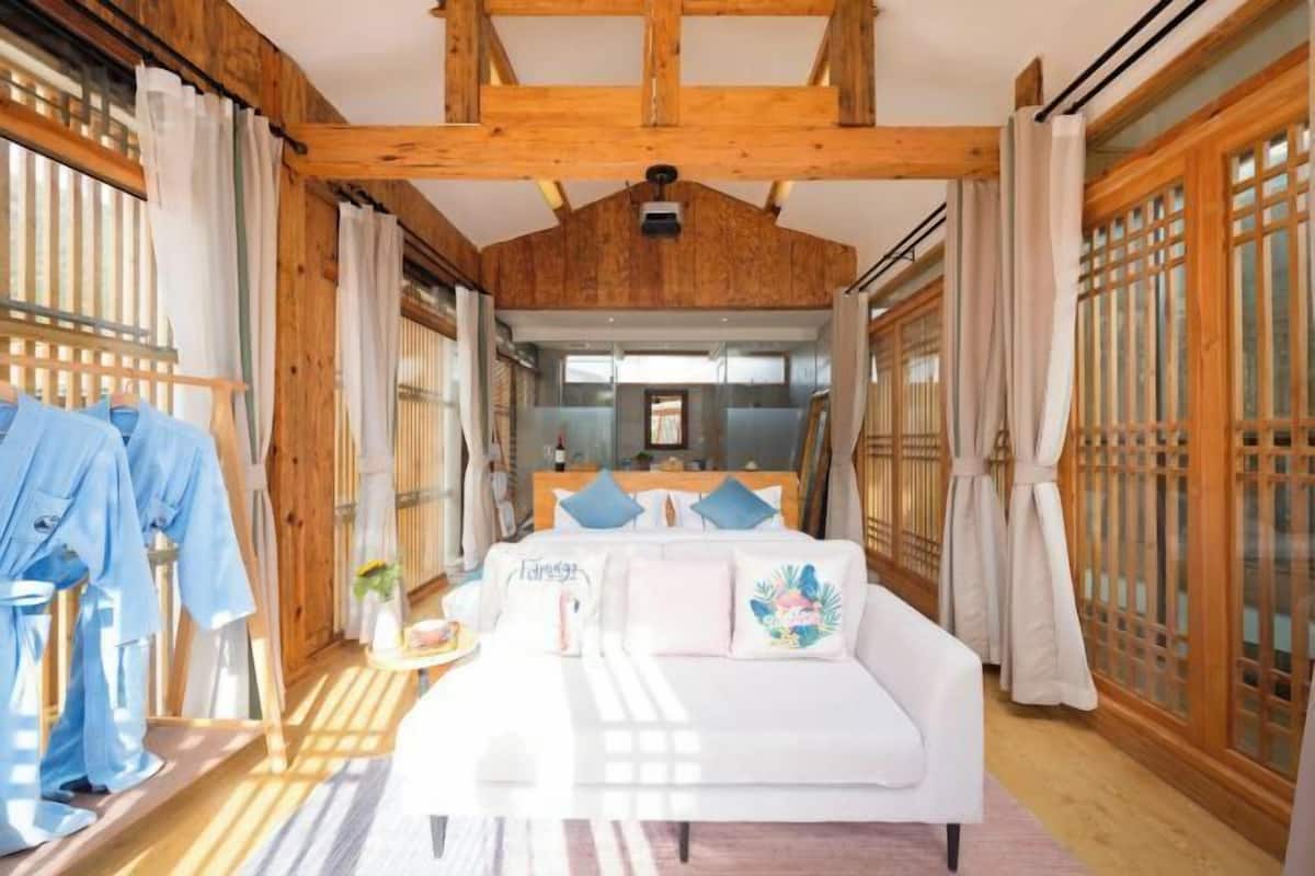 Una habitación con techo de madera, una cama con almohadas, una mesita con un jarrón y una ventana con cortinas.