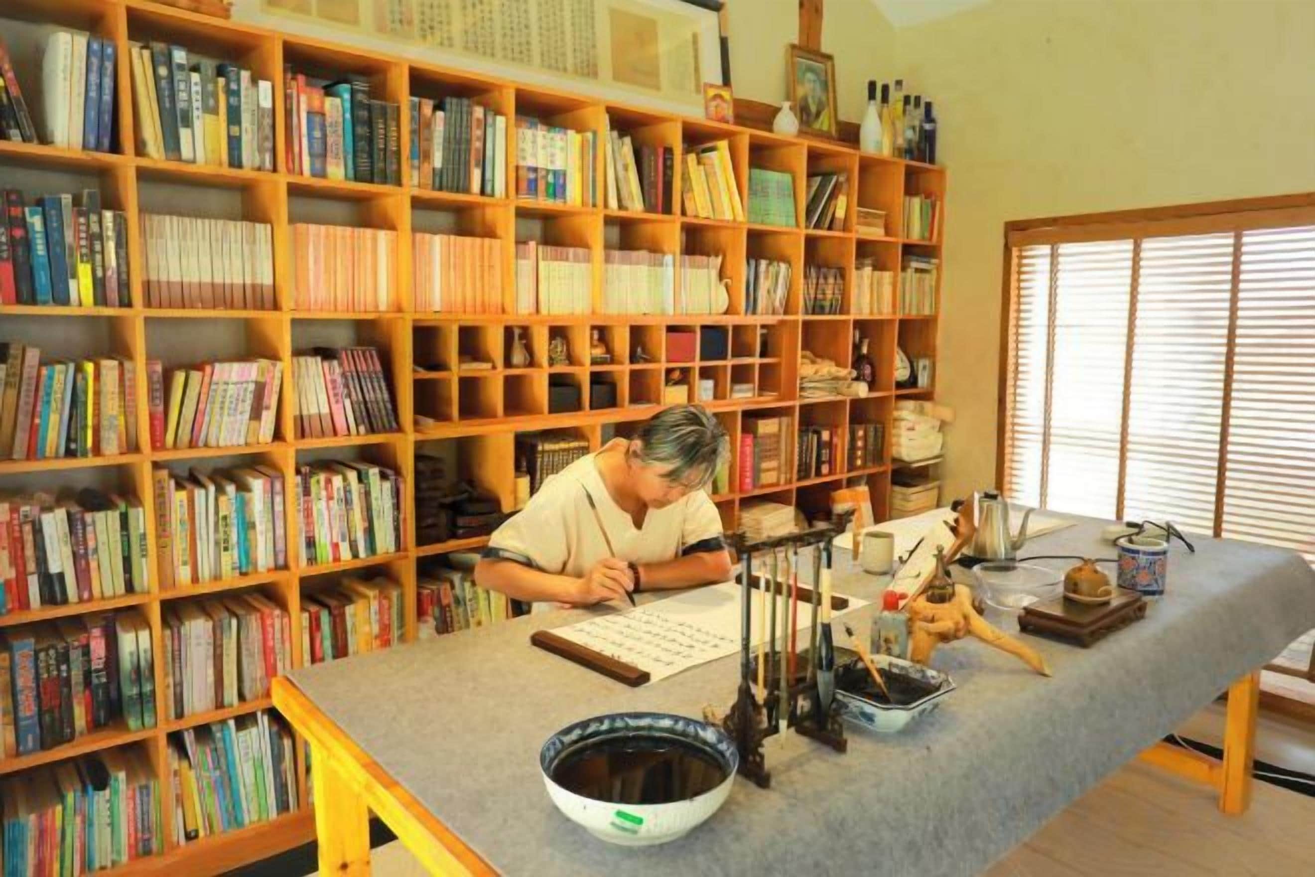 A room with a large bookshelf filled with books, a drafting table with drawing tools, and a window with blinds.