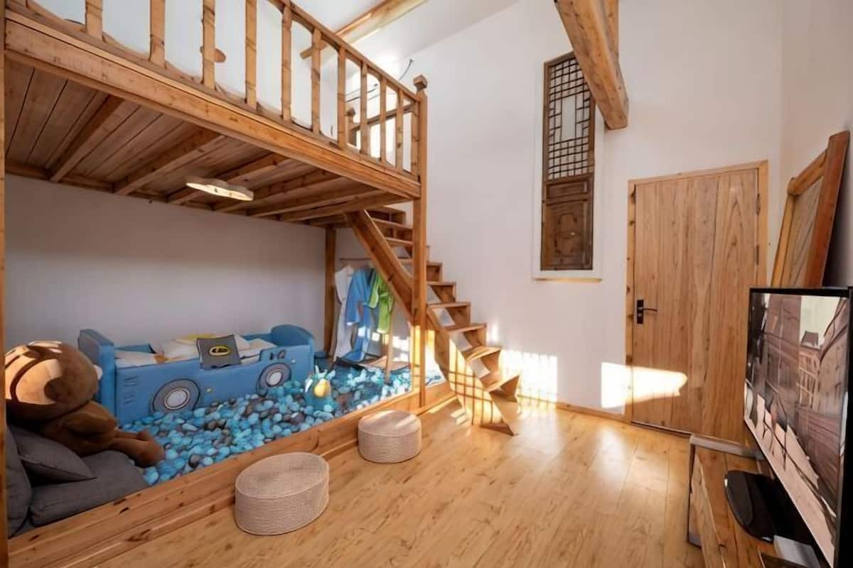 Una habitación con cama alta, un área de juegos con un tren azul y pelotas, una escalera de madera y un televisor.