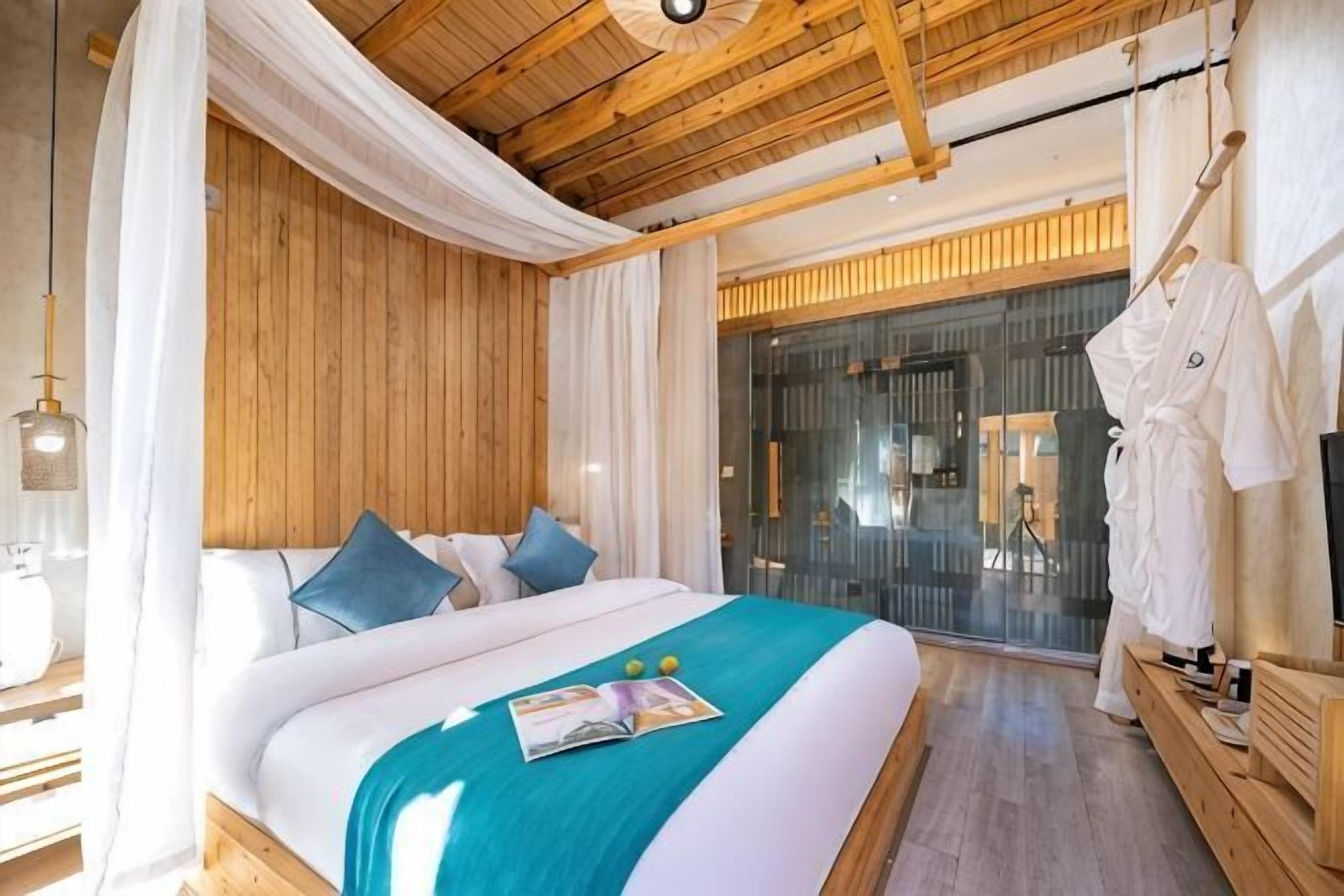 A bedroom with a wooden ceiling, a bed with white and teal bedding, a magazine on the bed, and a robe hanging on a rack.