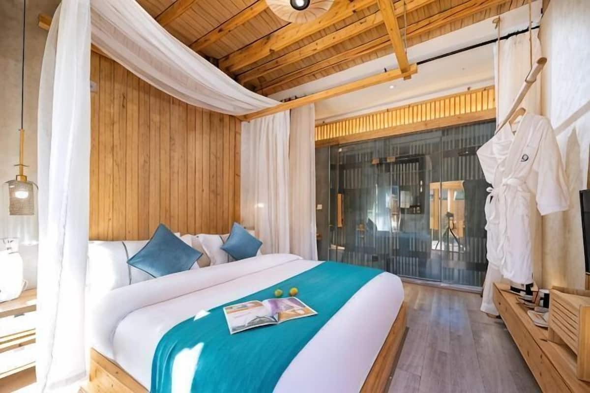 Un dormitorio con techo de madera, cama con ropa de cama blanca y turquesa, una revista sobre la cama y una bata colgada en un perchero.