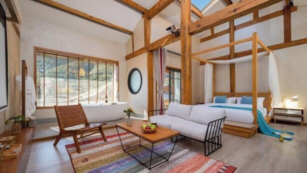 Ein geräumiges Zimmer mit hohen Decken, Holzbalken, einem Bett, einem Sofa, einem Sessel, einem Tisch und einem großen Fenster mit Blick.