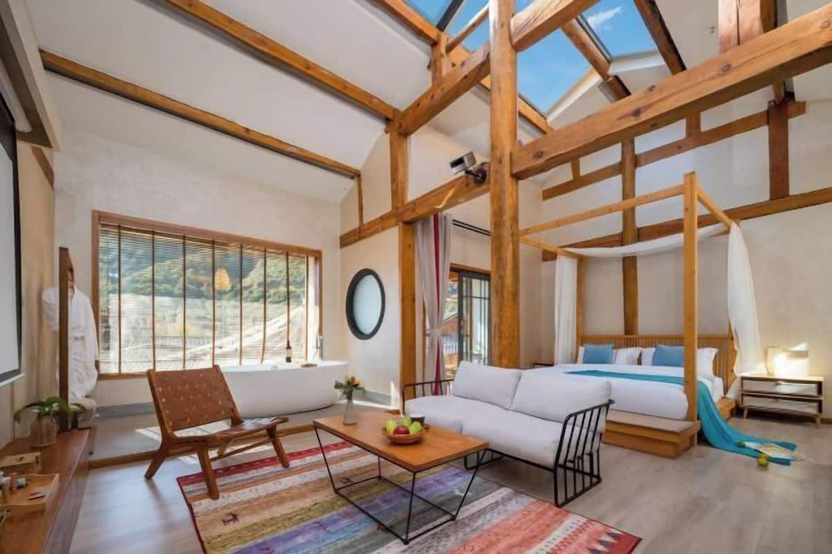 Amplia habitación con techo alto, vigas de madera, una cama, un sofá, una silla, una mesa y un gran ventanal con vistas.