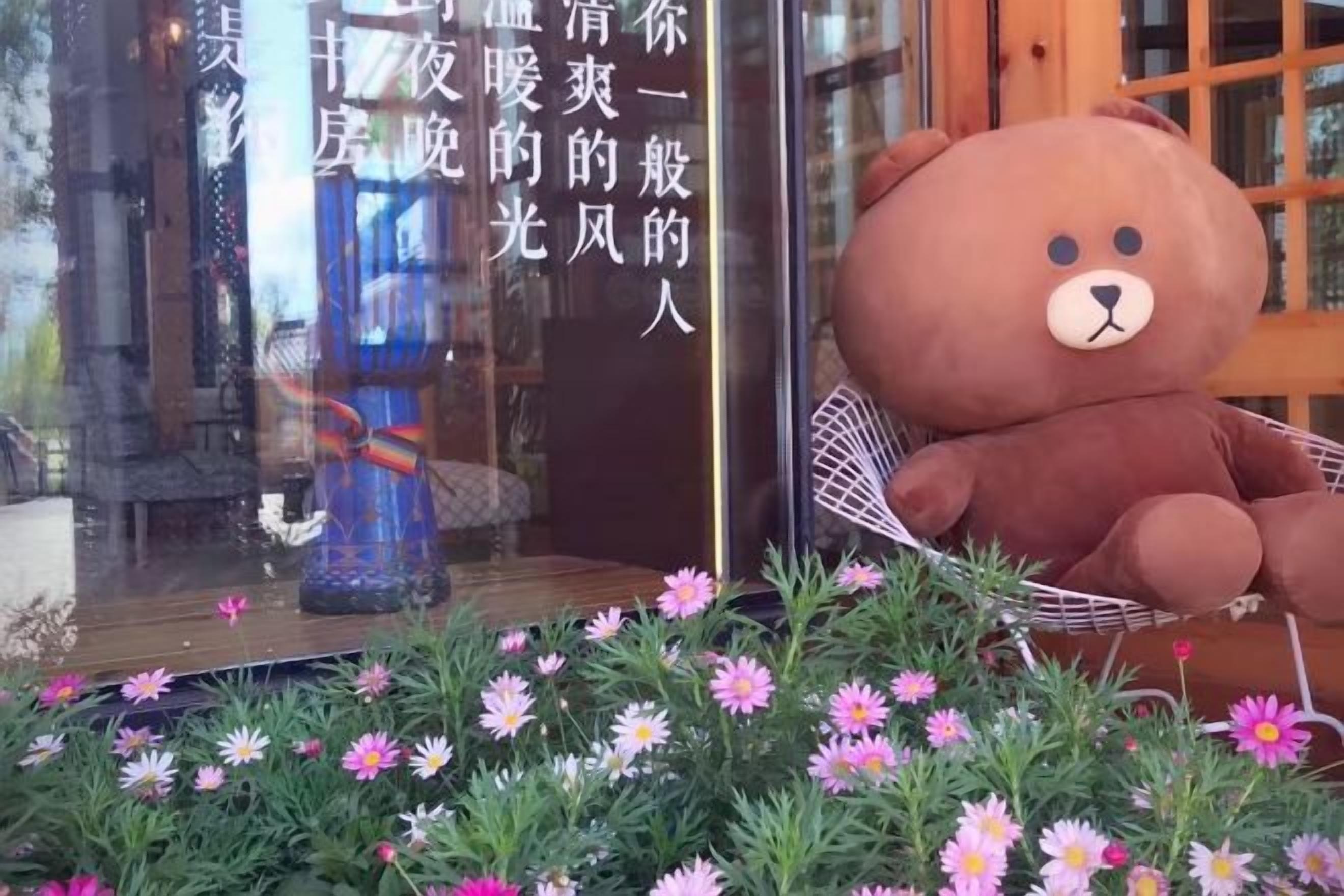 Ein großer brauner Teddybär sitzt in einem Korbstuhl, auf einer Holztischplatte mit einer Blumenkomposition davor.