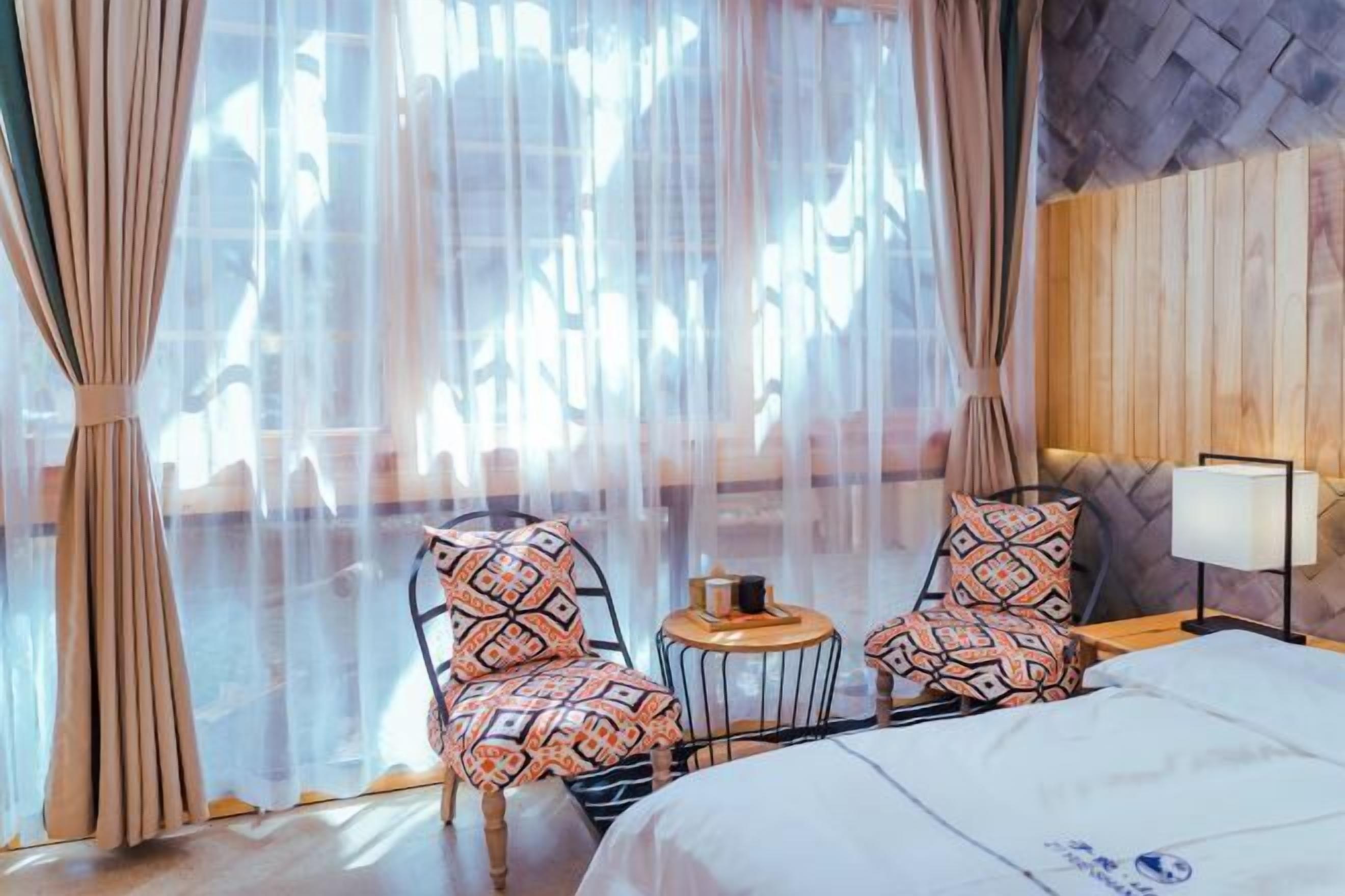 Una habitación de hotel con una cama, dos sillas con cojines estampados, una mesita redonda y una lámpara.