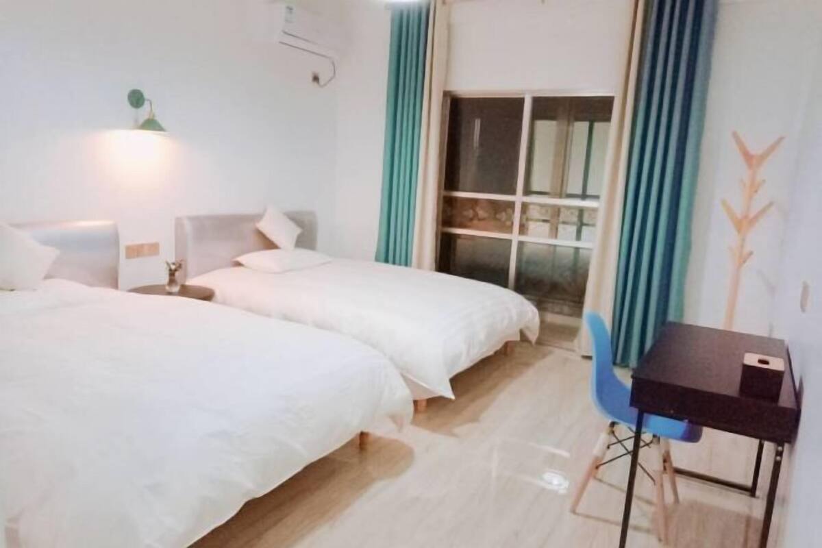 Habitación de hotel con dos camas, un escritorio, una silla, ventana con cortinas y una lámpara de pared.