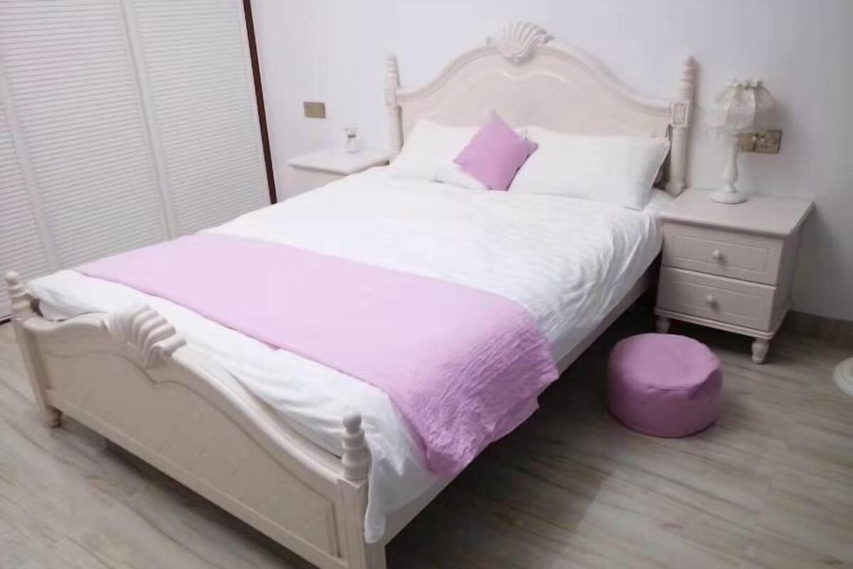 Un dormitorio con cama, mesitas de noche, cajón y un pouf redondo.