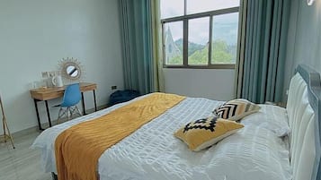 A bedroom with a bed, a desk, a chair, a window with curtains, and a view of greenery outside.