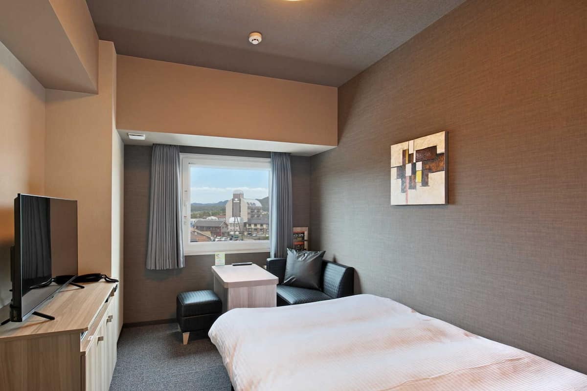 Habitación de hotel con cama, escritorio, silla, televisor y ventana con vista a la ciudad.