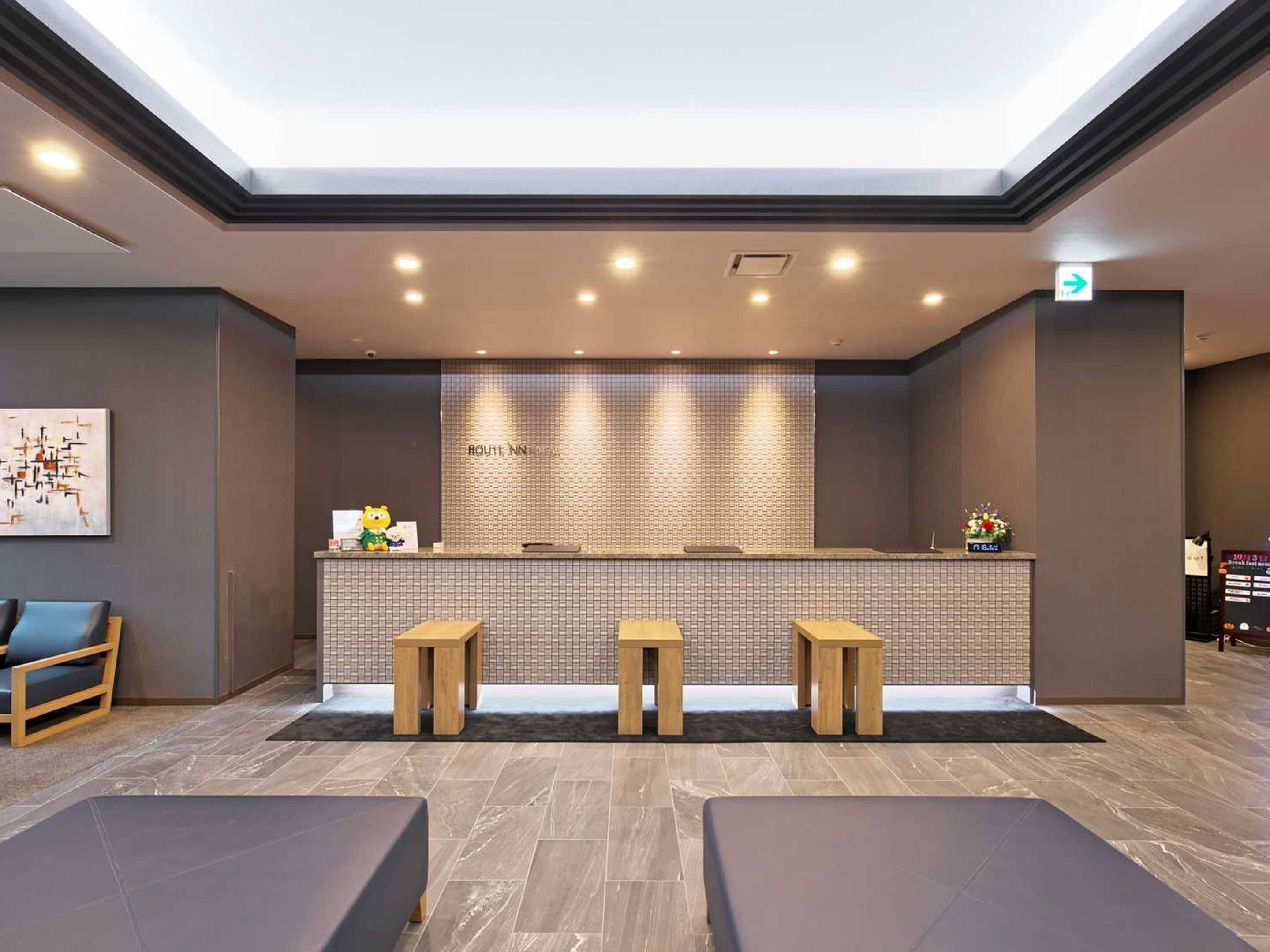 A reception area with a modern design, featuring a reception desk, three wooden stools, a seating area with two sofas, and a tiled wall with the name 'ROUTE NISSAN'.