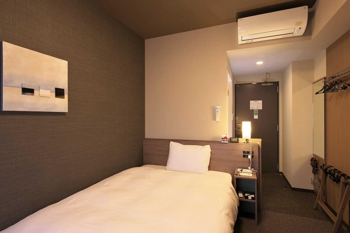 Una habitación de hotel con cama, aire acondicionado, mesita de noche con lámpara, espejo y perchero.