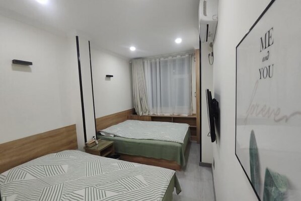 A hotel room with two beds, a window with sheer curtains, a wall-mounted TV, and a poster with the text "ME and YOU forever".