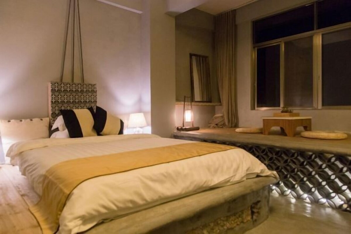 A hotel room with a large bed, a small bedside table, a window with curtains, and a hanging light fixture.