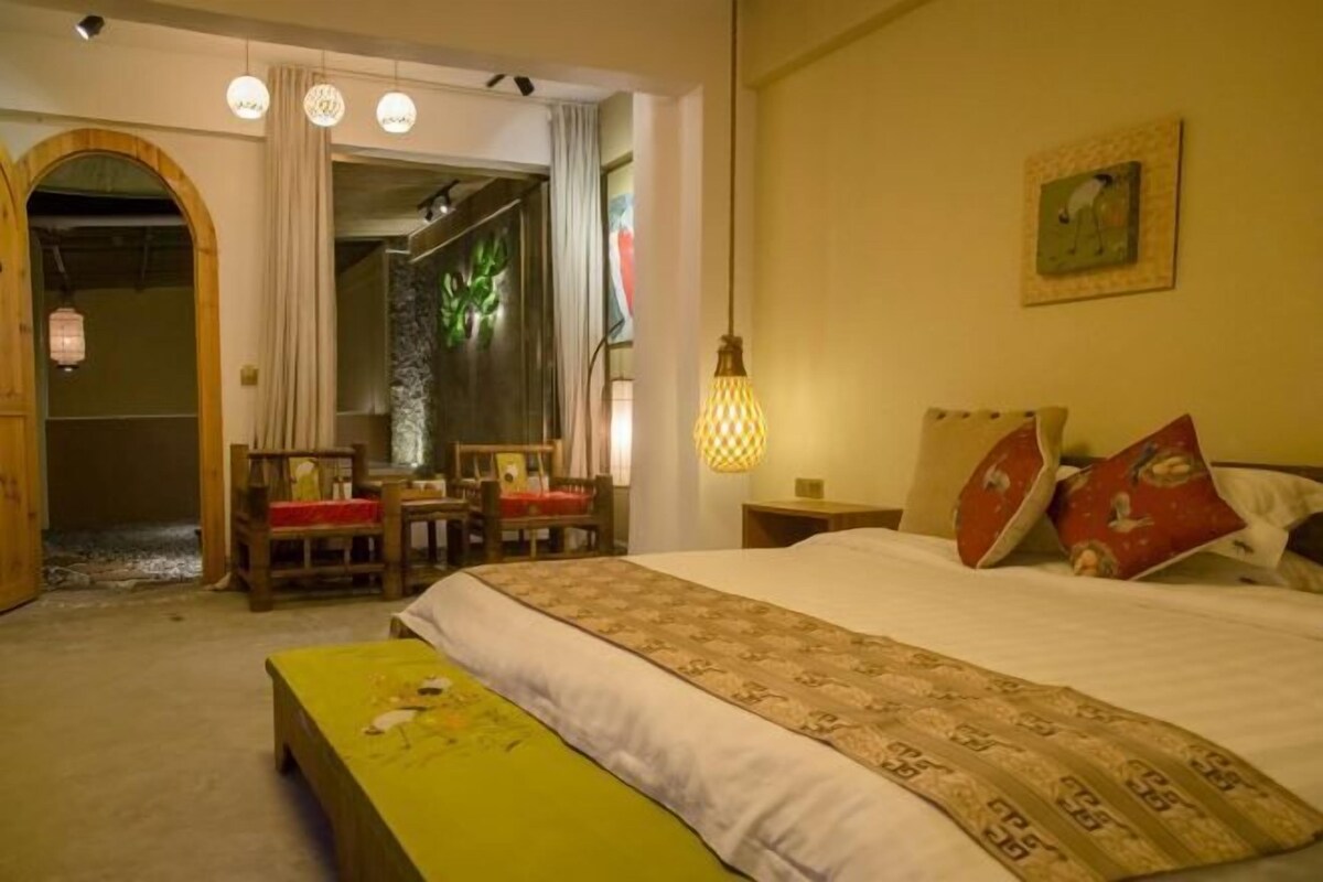 A hotel room with a bed, a bench, a desk, and a chair. There is a window with curtains and a painting on the wall.