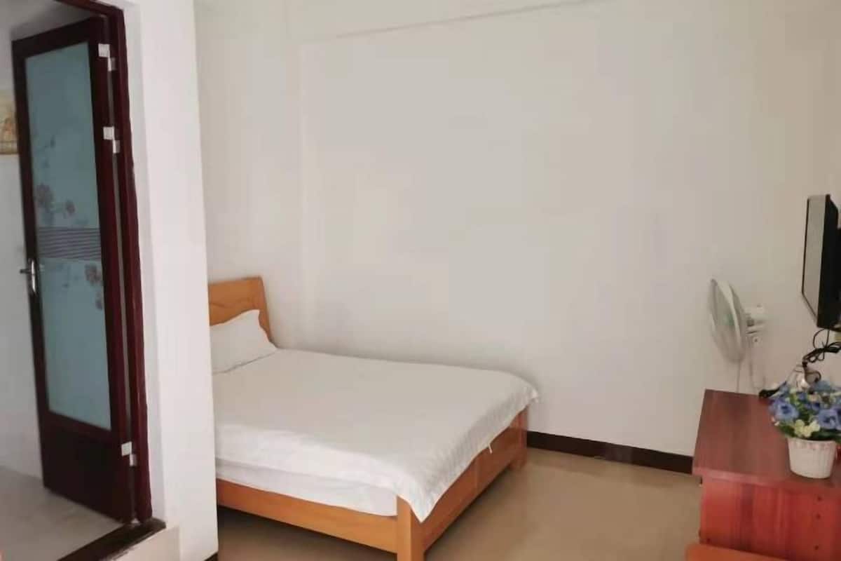 A single bed in a room with a wooden headboard, a white wall, a door with a floral design, and a small table with a potted plant.