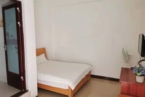 A single bed in a room with a wooden headboard, a white wall, a door with a floral design, and a small table with a potted plant.