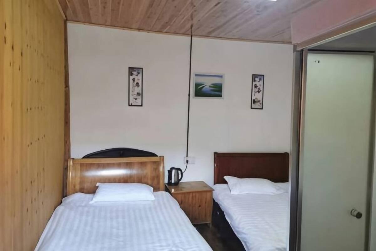 A room with two beds, wooden headboards, a nightstand with a kettle, and a door.