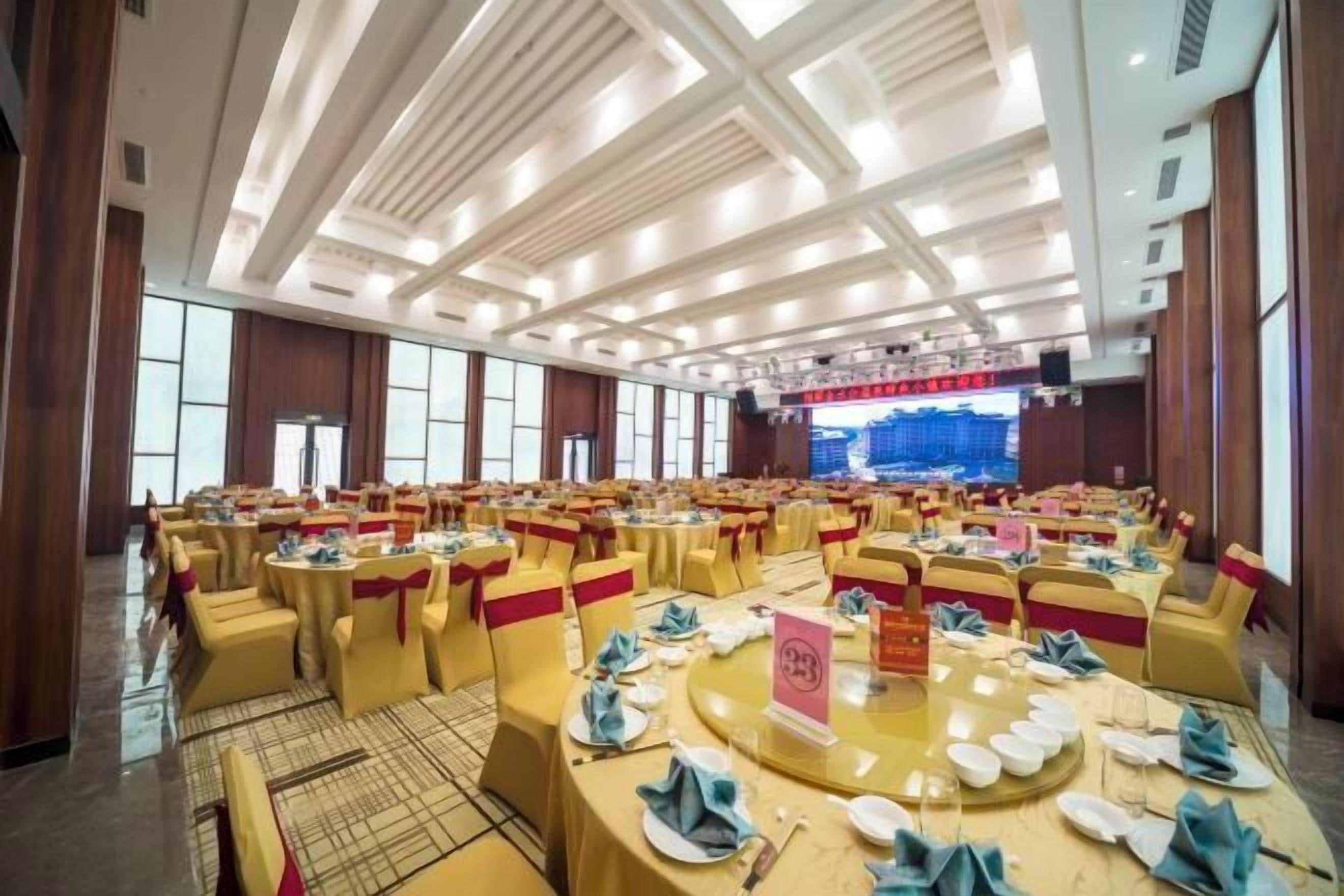 A banquet hall with round tables, yellow chairs, and a stage with a large screen.