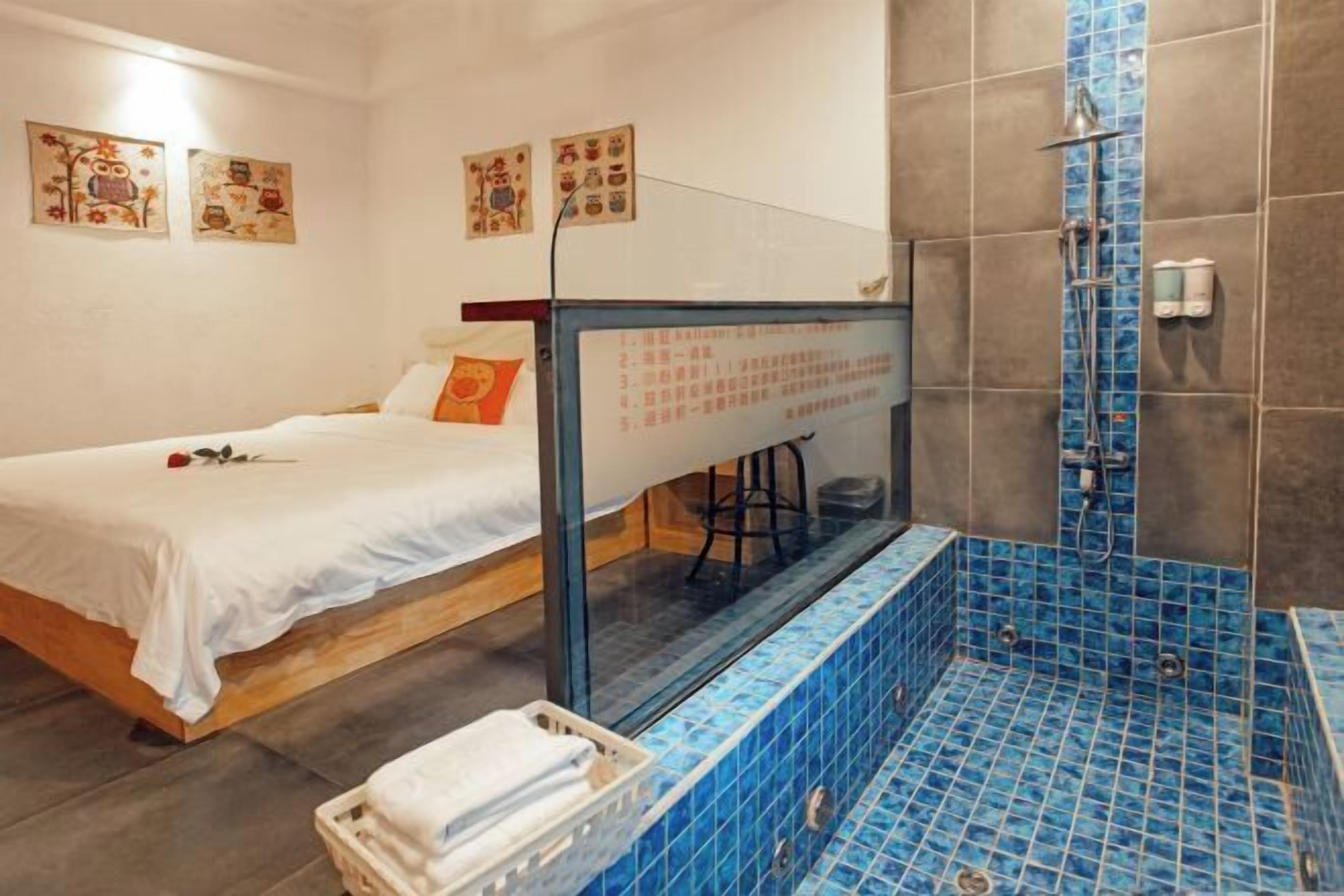 A hotel room with a bed, a small stool, a shower area with blue tiles, and artwork on the walls.