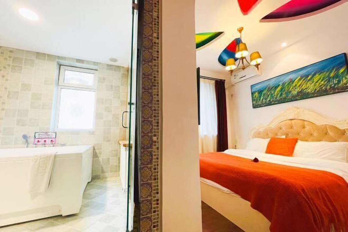 A hotel room with a bathroom, a bed with an orange bedspread, and a colorful wall painting.