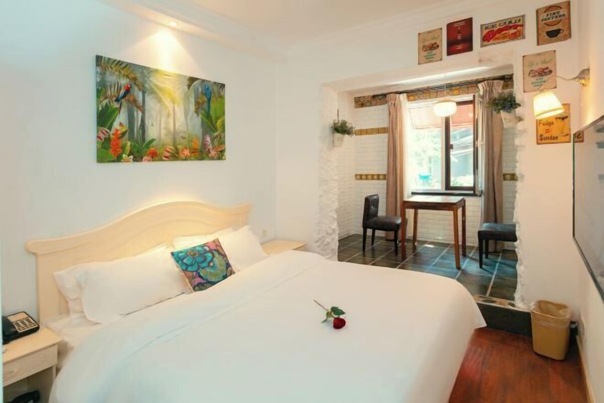 A hotel room with a bed, a desk, and a chair. There is a painting on the wall and a window with curtains.