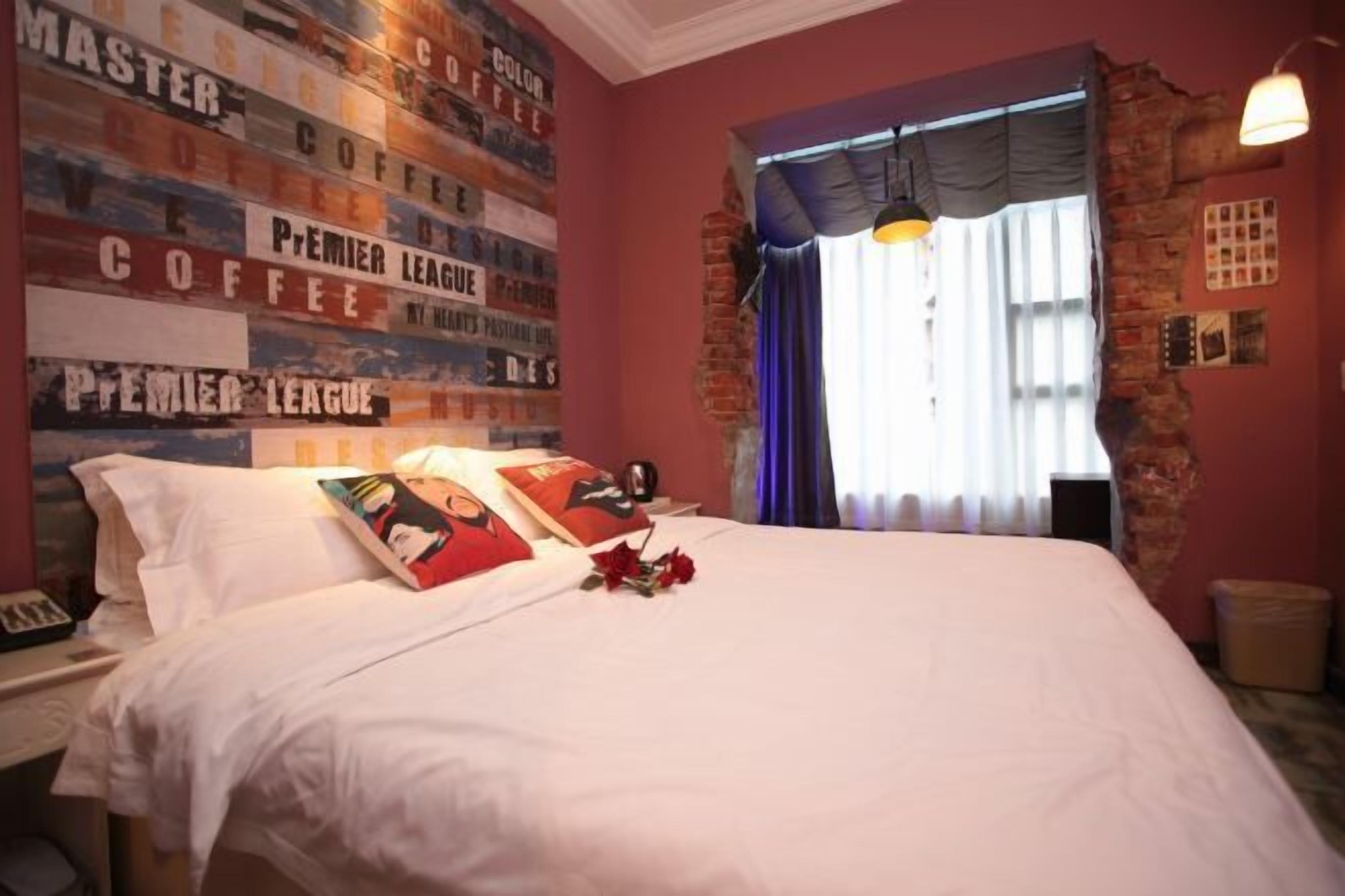 A hotel room with a bed, a wall with coffee-related posters, a window with curtains, and a brick wall.