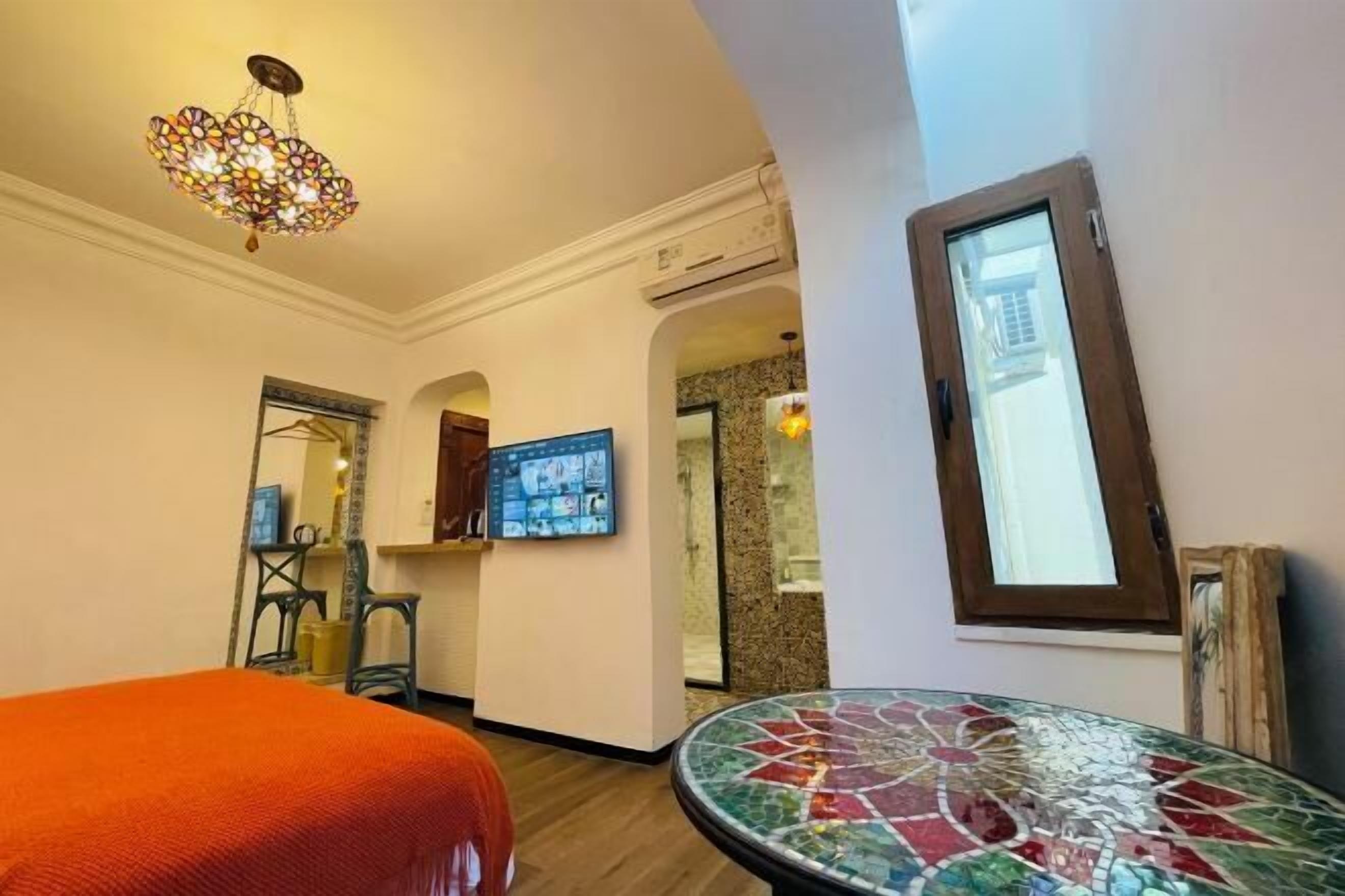 Una habitación de hotel con cama, una mesita de mosaico, un televisor montado en la pared y un candelabro decorativo.