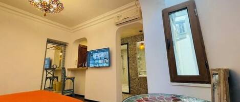 Una habitación de hotel con cama, una mesita de mosaico, un televisor montado en la pared y un candelabro decorativo.