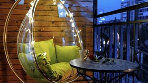 Una silla colgante con cojines y luces de cadena, una mesita redonda pequeña con una taza y un libro, y una barandilla de balcón.