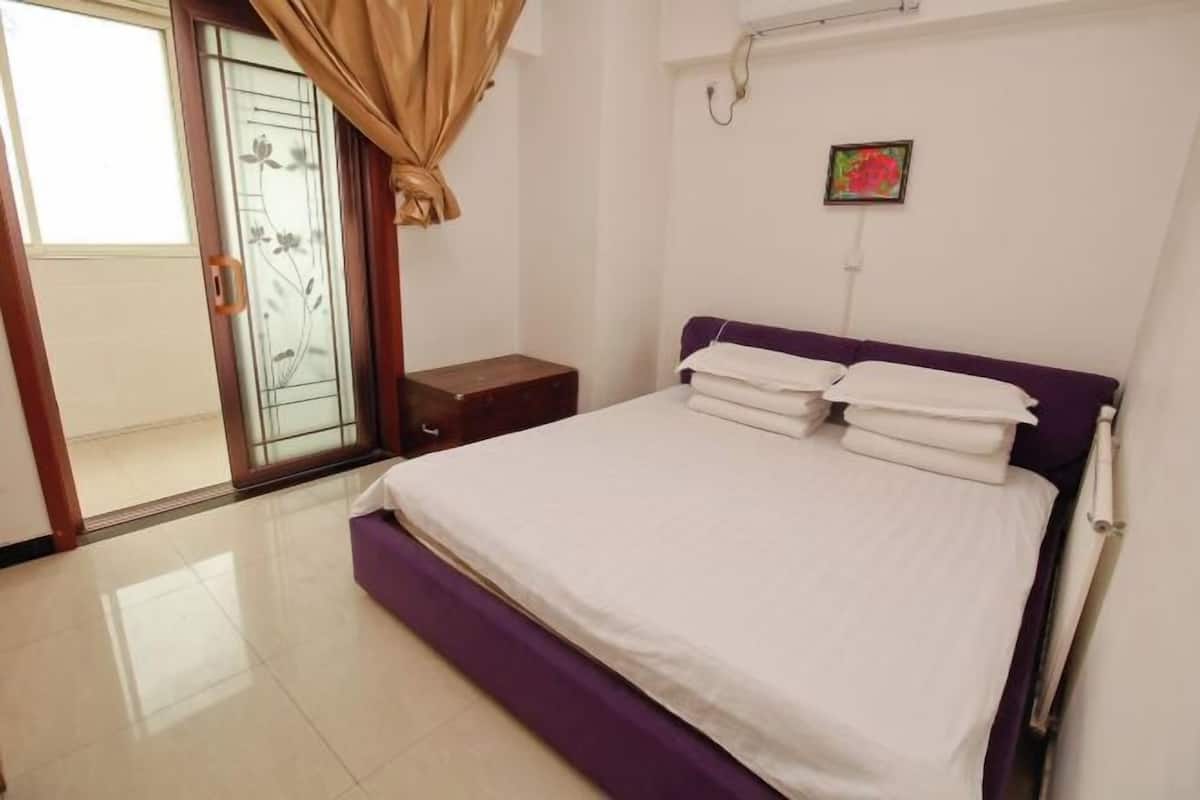 A bedroom with a bed, a nightstand, a window with curtains, and a small painting on the wall.