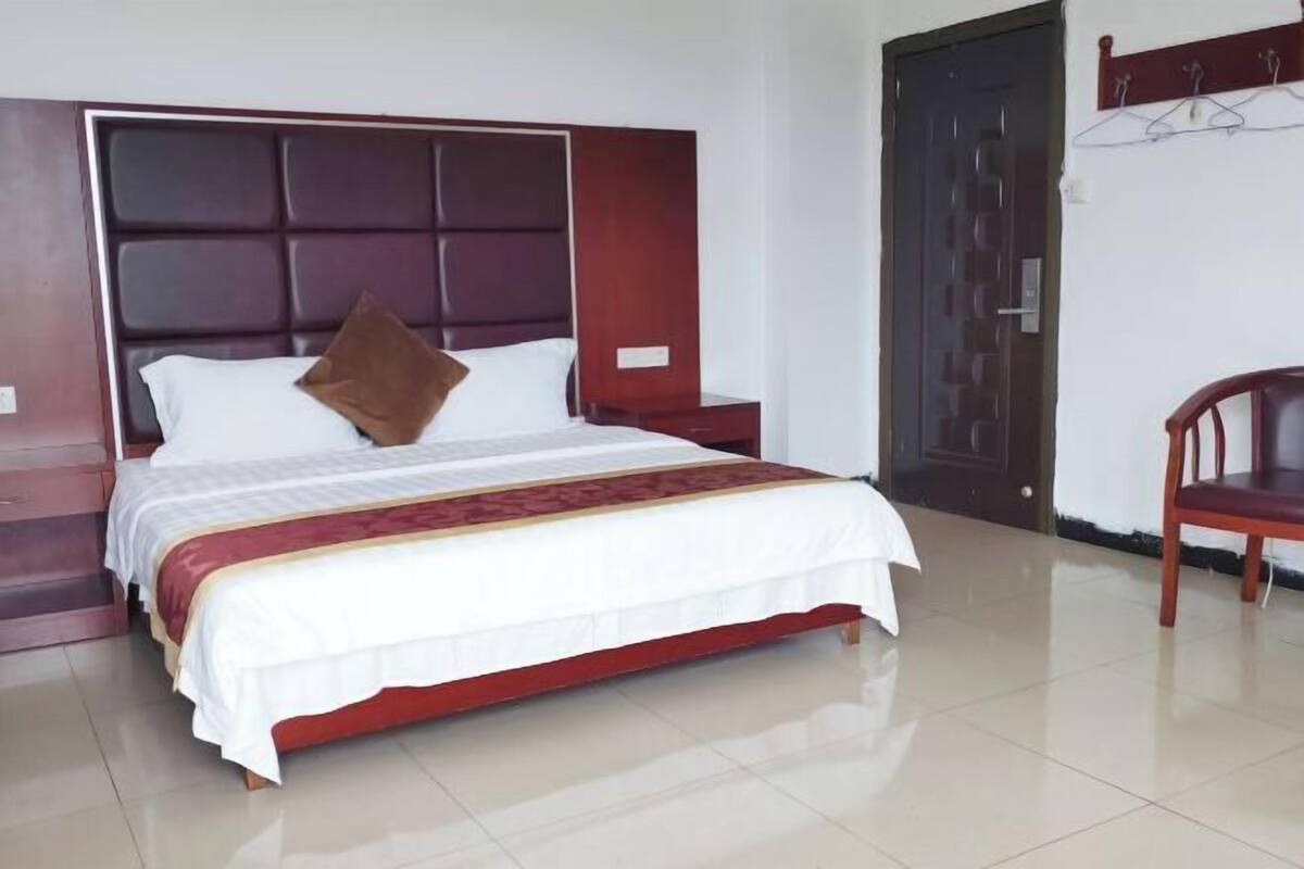 Een opgemaakt bed met hoofdeinde, een houten stoel en een deur in een kamer met betegelde vloer.