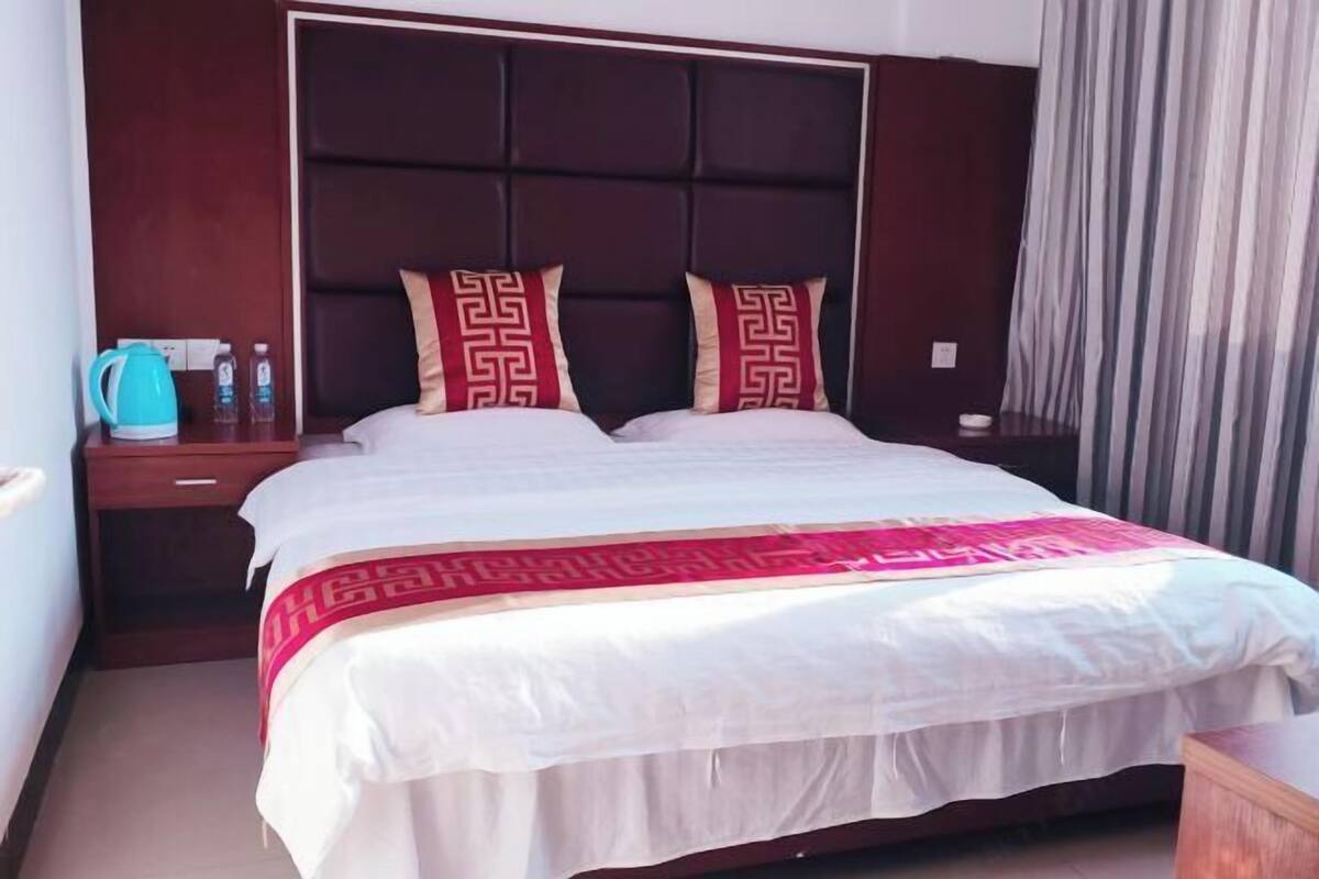 Een hotelkamer met een groot bed, twee kussens met een geometrisch patroon, een nachtkastje met een waterkoker en waterflessen, en een gordijn voor het raam.