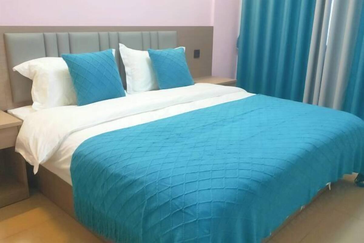 Een bed met een blauwe quilt, witte kussens, een grijs hoofdbord en een blauw gordijn op de achtergrond.