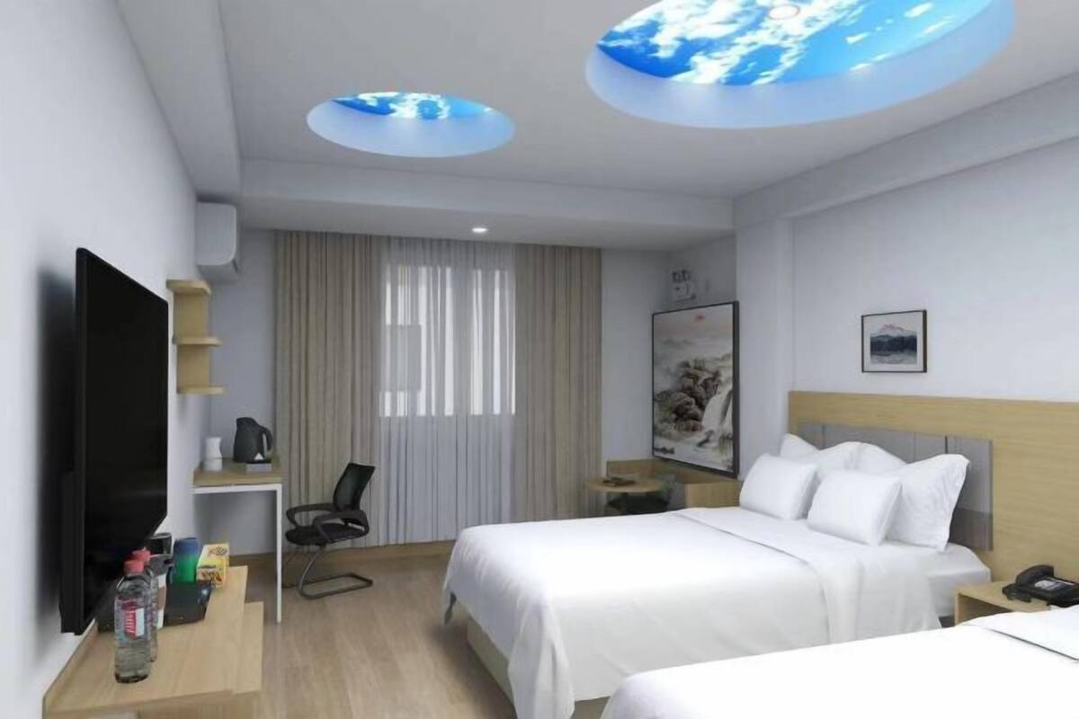 Habitación de hotel con dos camas, un televisor de pantalla plana, un escritorio con una silla, una cafetera y una lámpara circular en el techo.