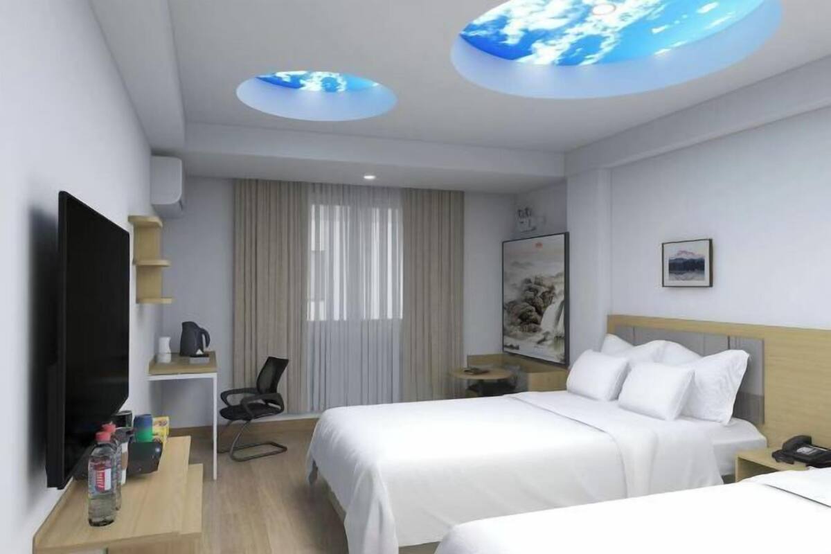 Habitación de hotel con dos camas, un televisor de pantalla plana, un escritorio con silla, cafetera y lámpara circular en el techo.